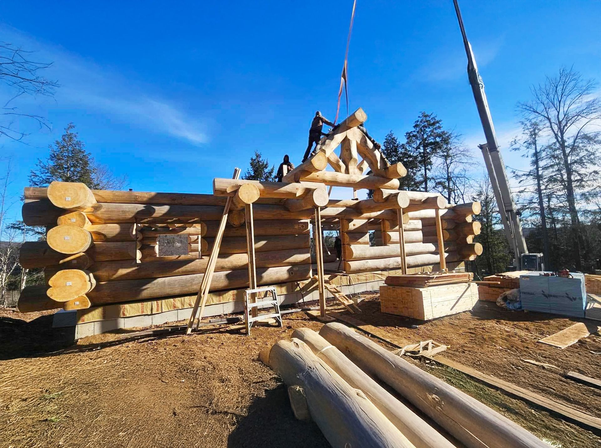 Workers assembling a log cabin frame with crane, stacked logs, and snowy winter ground under a blue sky