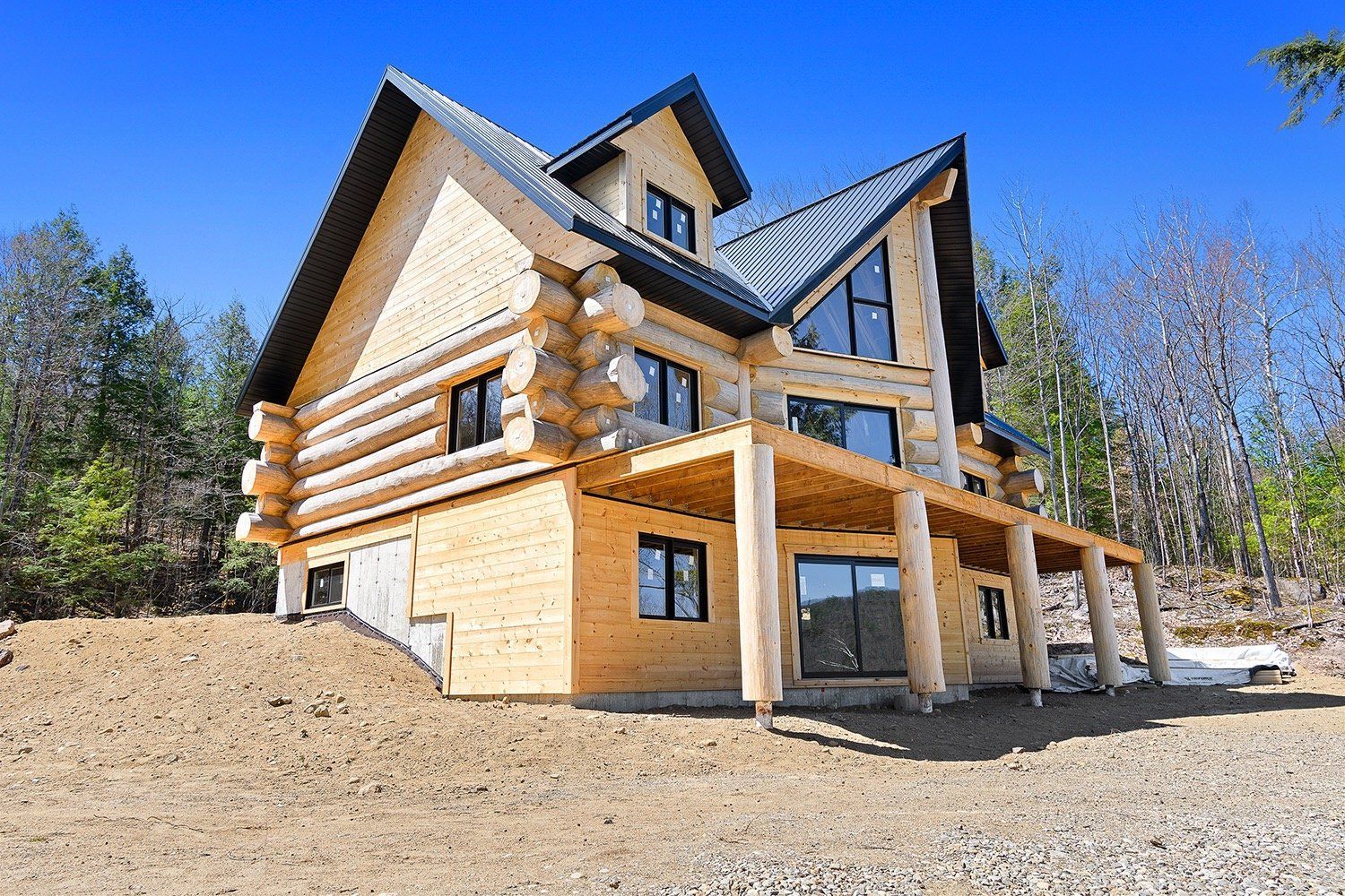 Large log cabin with a covered porch on a sunny hillside under a clear blue sky
