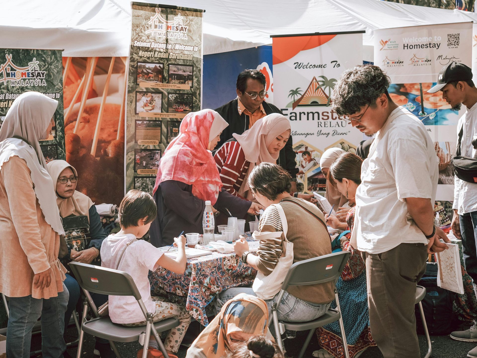 People gathered around a table at an outdoor market booth, with tents and banners in the background.