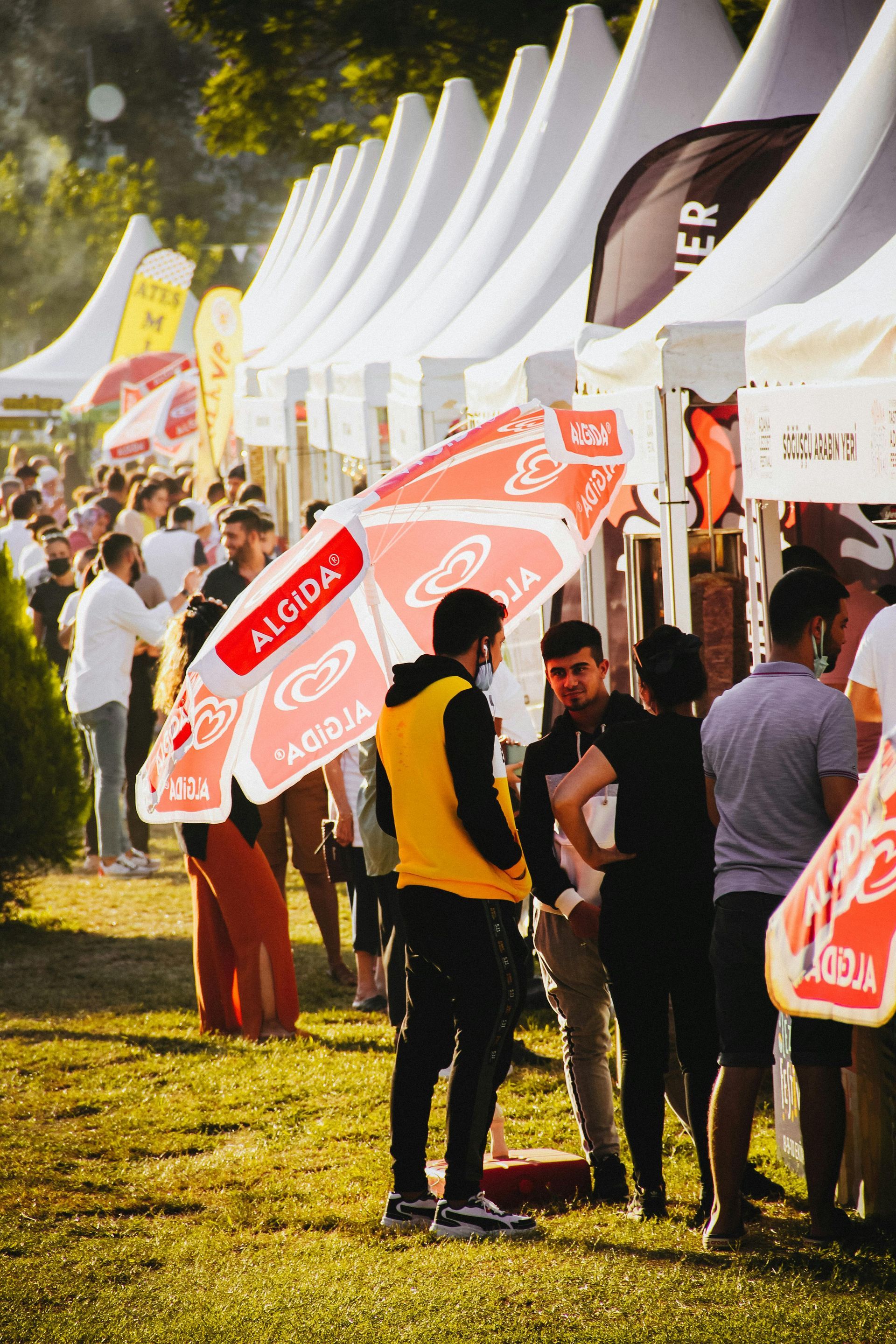 People holding red signs in a sunny outdoor event queue beside white tents and flags.