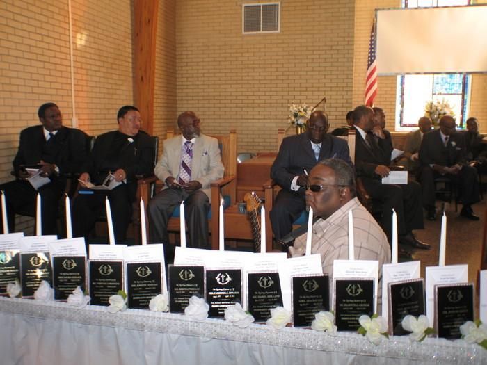 Several men seated indoors behind a table lined with black-and-white awards or programs