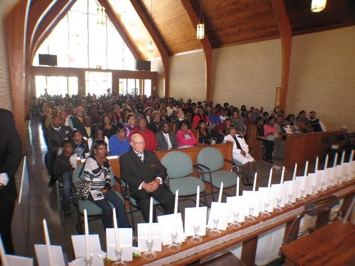 Audience seated in a church hall with wood-paneled ceiling and rows of white candles in front