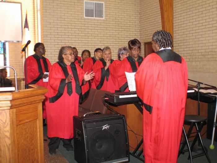 Choir in red robes singing indoors beside a keyboard and speaker