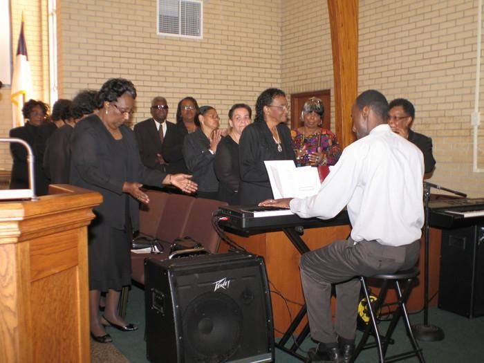Choir singing beside a pianist at a church service, with a speaker and lectern in the foreground