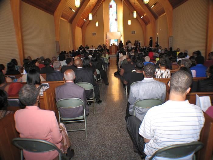 People seated in rows at a small church or hall, facing the front during a meeting or service.