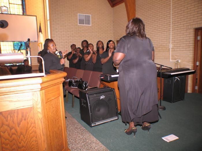 Choir singing in a church, with a woman leading beside a piano and speakers