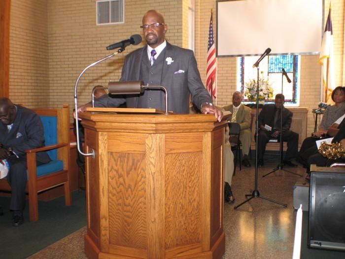 Man speaking at a wooden podium in a church, with musicians seated behind him and an American flag nearby