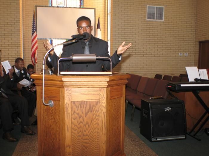 Speaker at a podium in a church, gesturing during a speech with chairs and a U.S. flag behind him