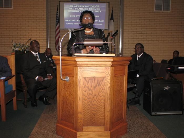 Woman speaking at a wooden podium in a church-like hall, with seated attendees behind her.