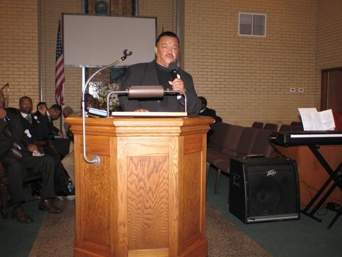 Man speaking at a wooden podium in a church or hall, with audience seated and keyboard nearby