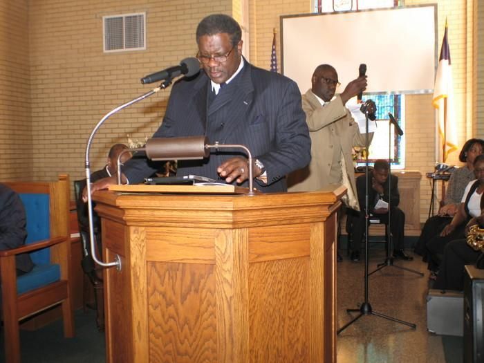 Man speaking at a wooden podium in a church, with a microphone and people seated in the background