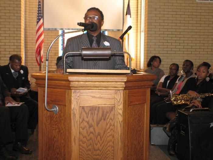 Person speaking at a podium in a church or meeting hall, with seated audience behind them