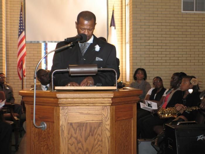 Man speaking at a wooden podium in a church, with seated audience and American flag in the background