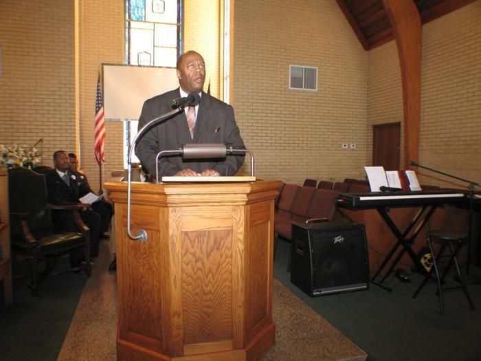 Man speaking at a wooden pulpit in a church, with a keyboard and chairs nearby.