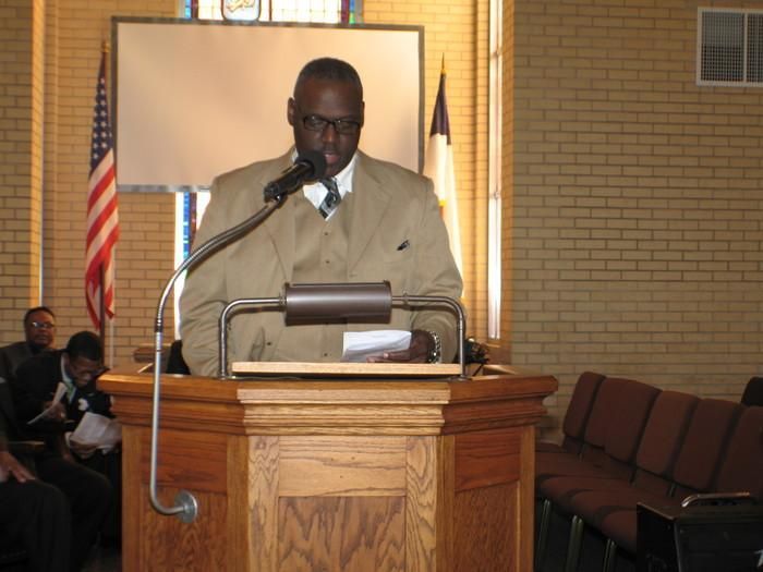 Man speaking at a podium in a church, with an American flag and empty chairs in the background