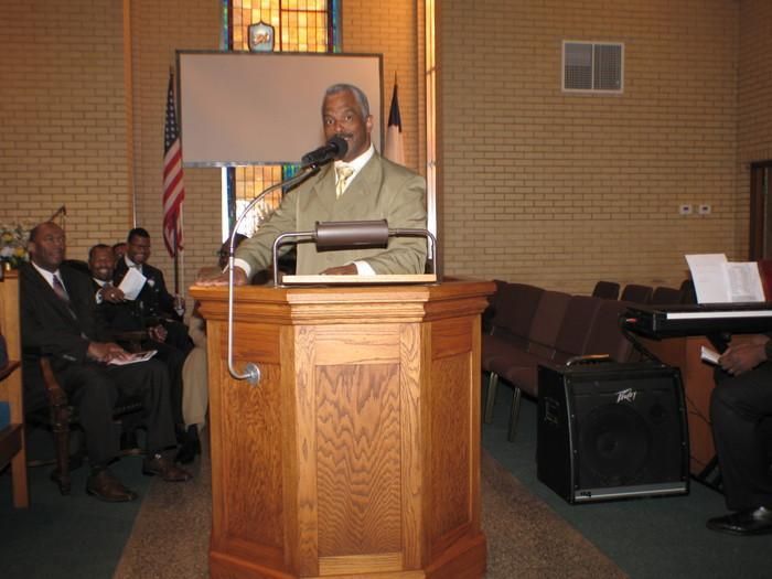 Man speaking at a podium in a church or meeting hall, with seated audience and American flag nearby