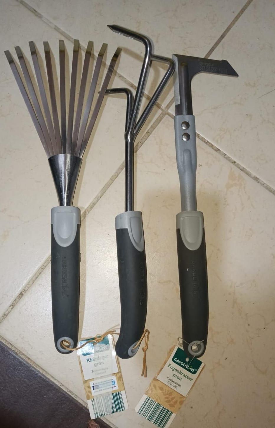 Three garden tools with black handles on a tiled floor: a fan rake, cultivator, and hoe-like scraper.