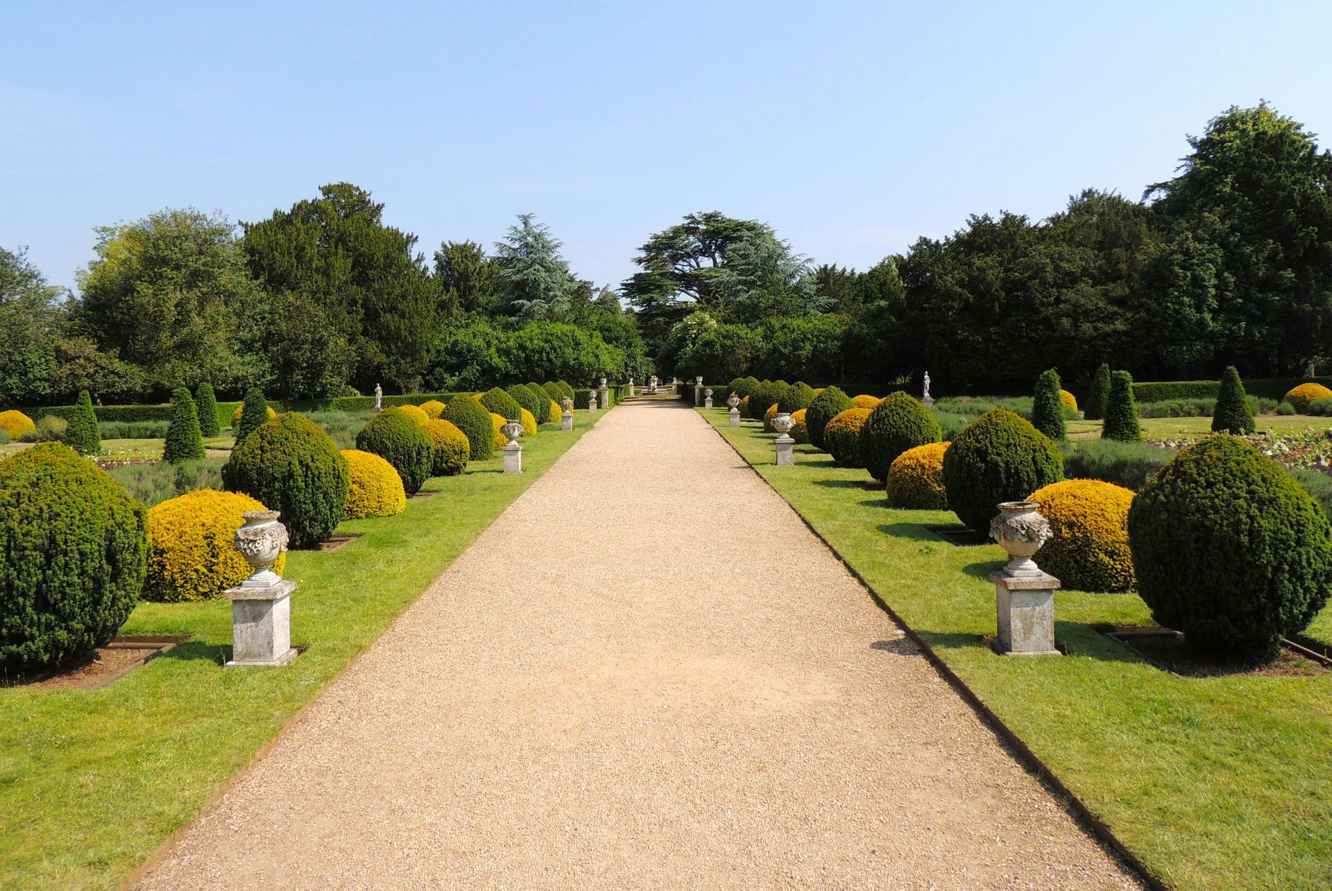 A gravel path leads through a formal garden lined with sculpted round bushes and ornamental stone urns on pedestals.