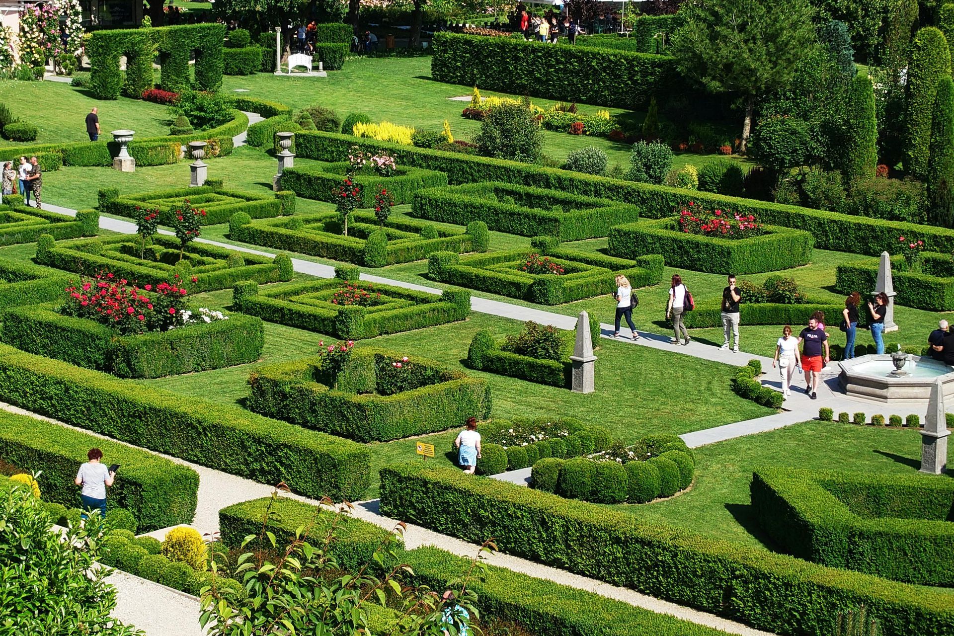 High-angle view of a manicured formal garden with geometric hedge patterns, flower beds, pathways, and visitors.