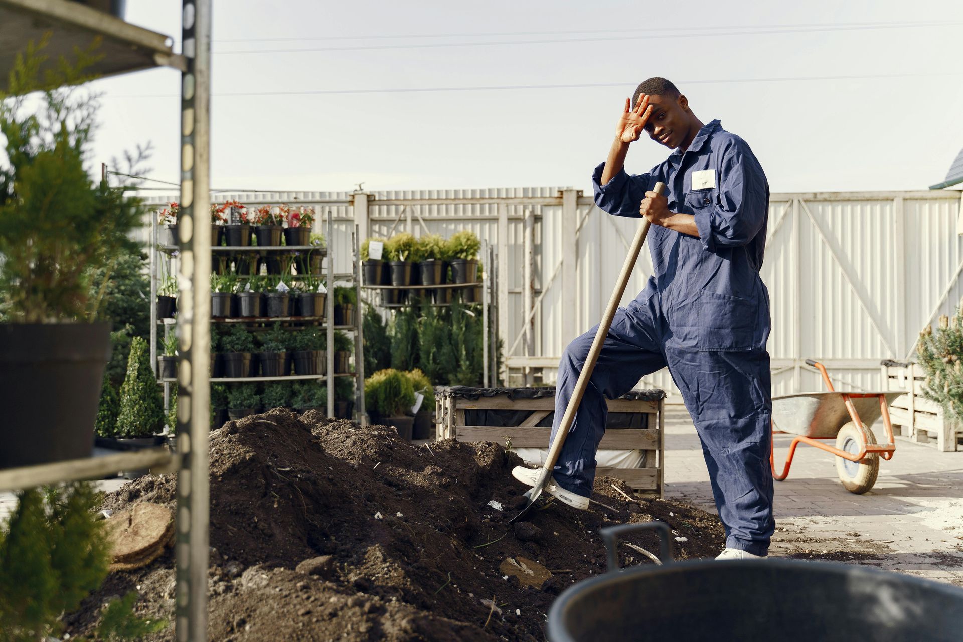 A person in blue coveralls takes a break from shoveling soil in a garden center, resting with a hand on their forehead.