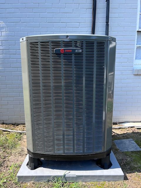 Gray Trane air conditioner unit on a concrete pad, against a white brick wall.