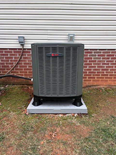 Dark green HVAC unit on a concrete pad with black supports, against a brick and siding wall.