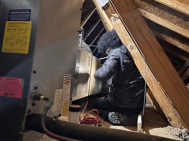 Person working on HVAC unit in an attic. They are wearing a jacket and kneeling.