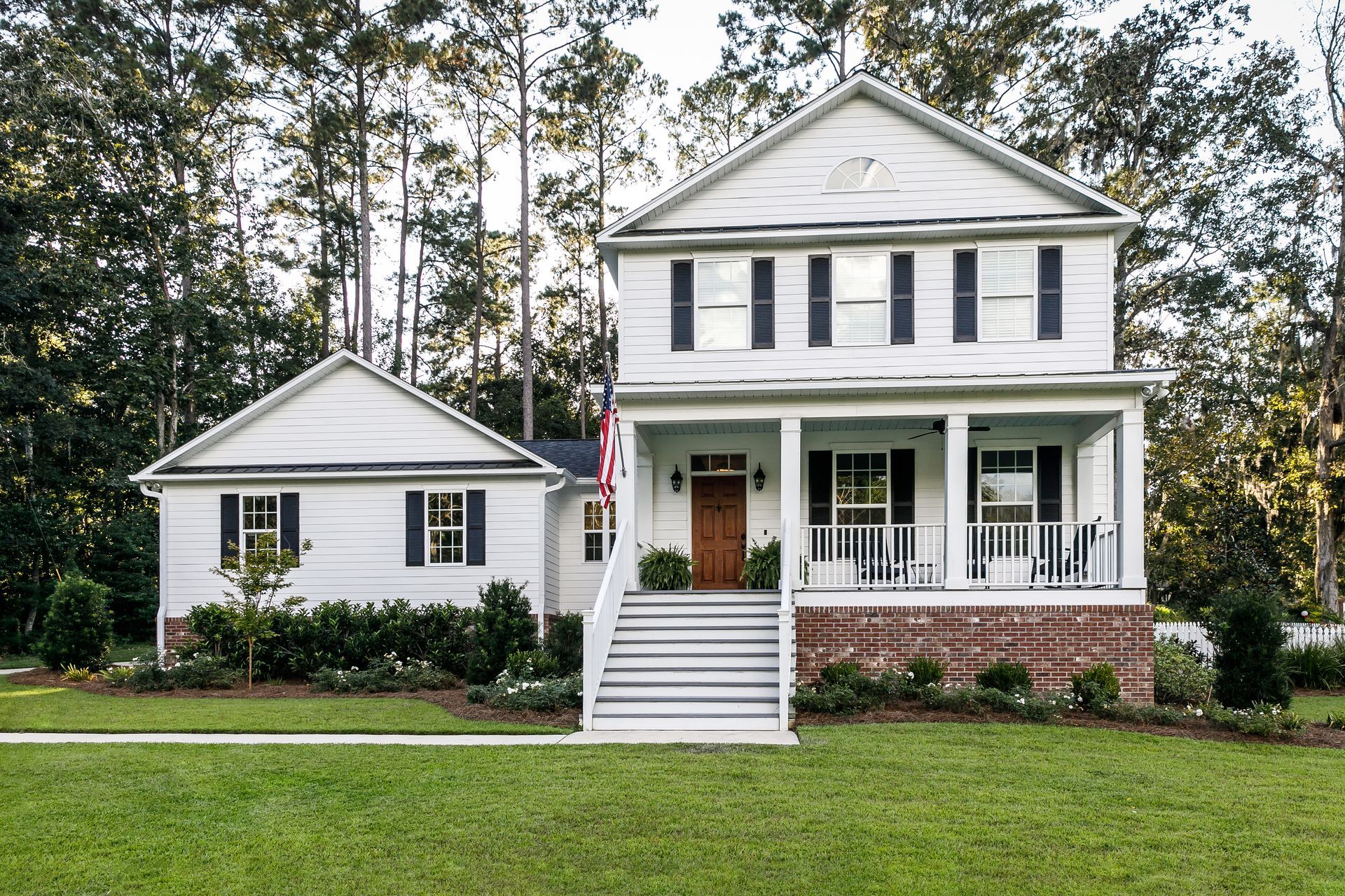 White two-story house with front porch, shutters, and attached small structure. Brick foundation, lawn, and trees.