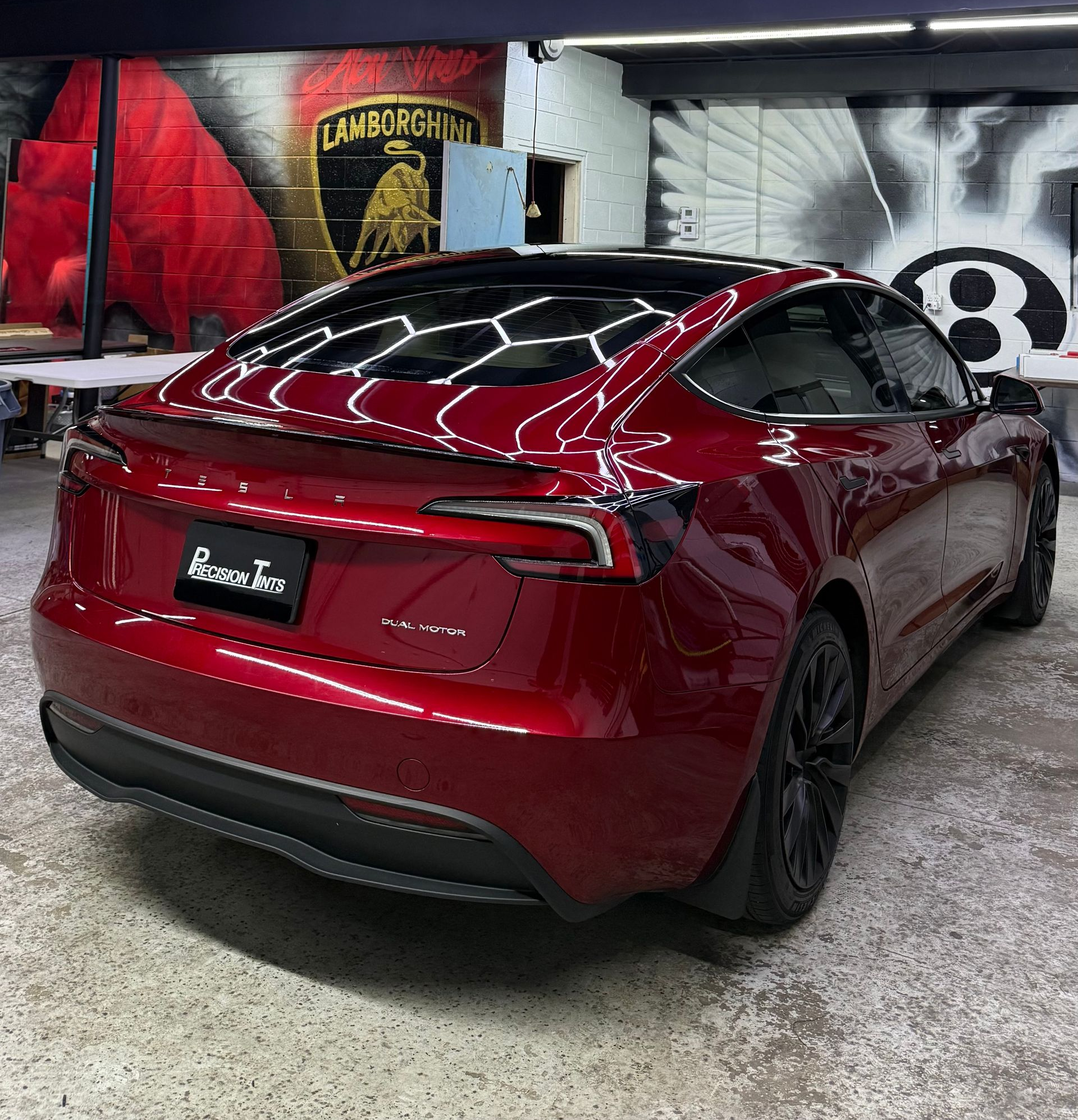 Red Tesla sedan parked indoors in a garage, shown from the rear-left against mural walls.
