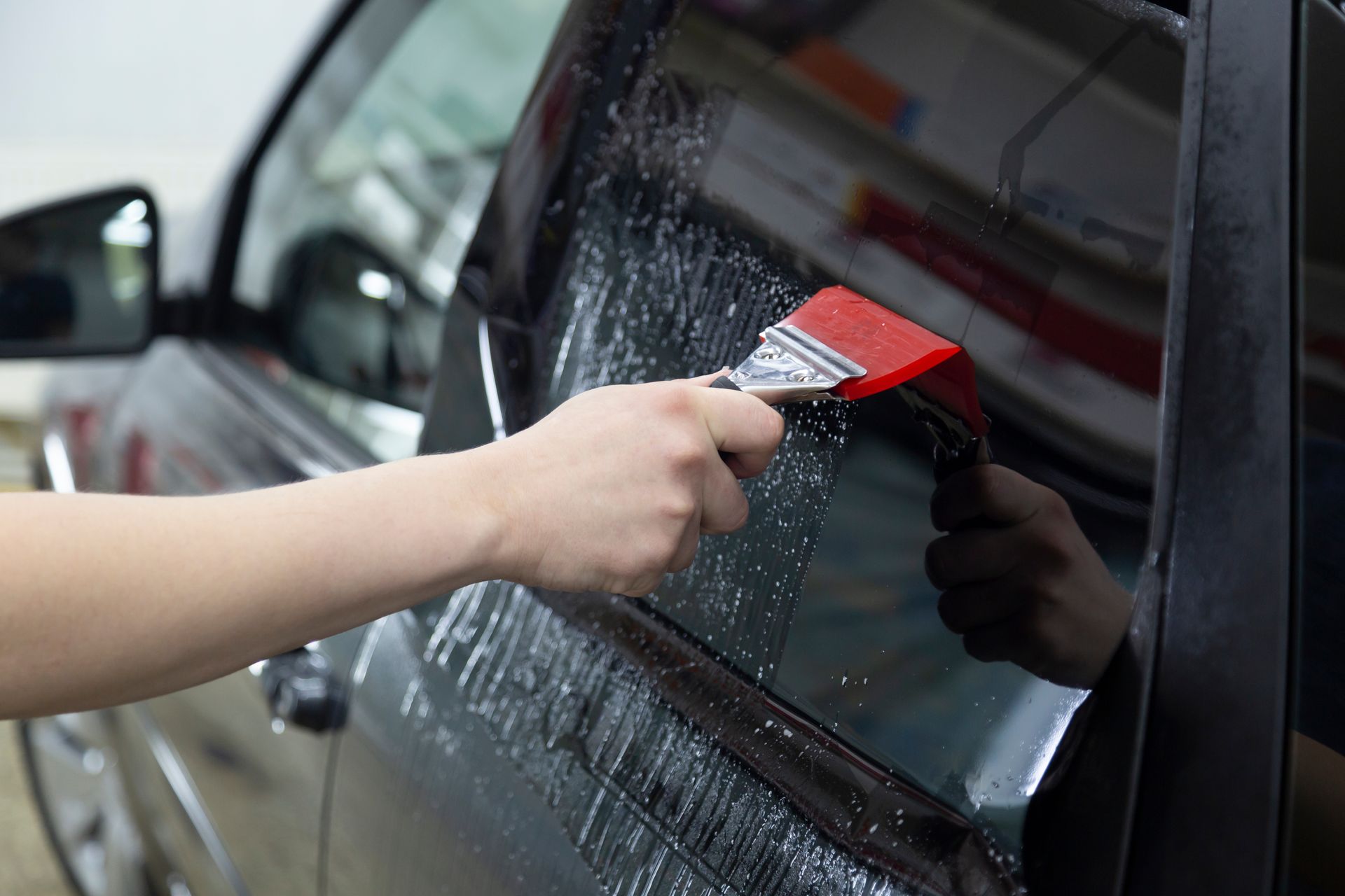 Hand cleaning a black car window with a red squeegee, removing water spots.
