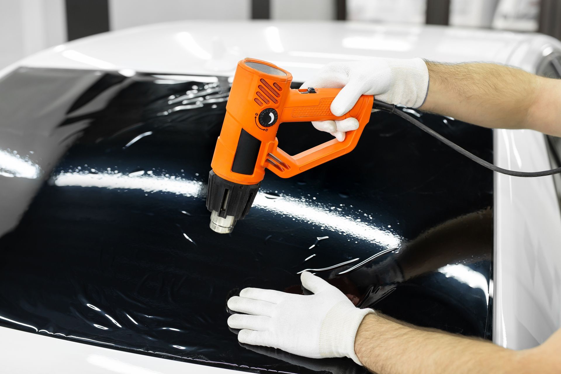 Gloved hands use an orange heat gun on a glossy black car hood.