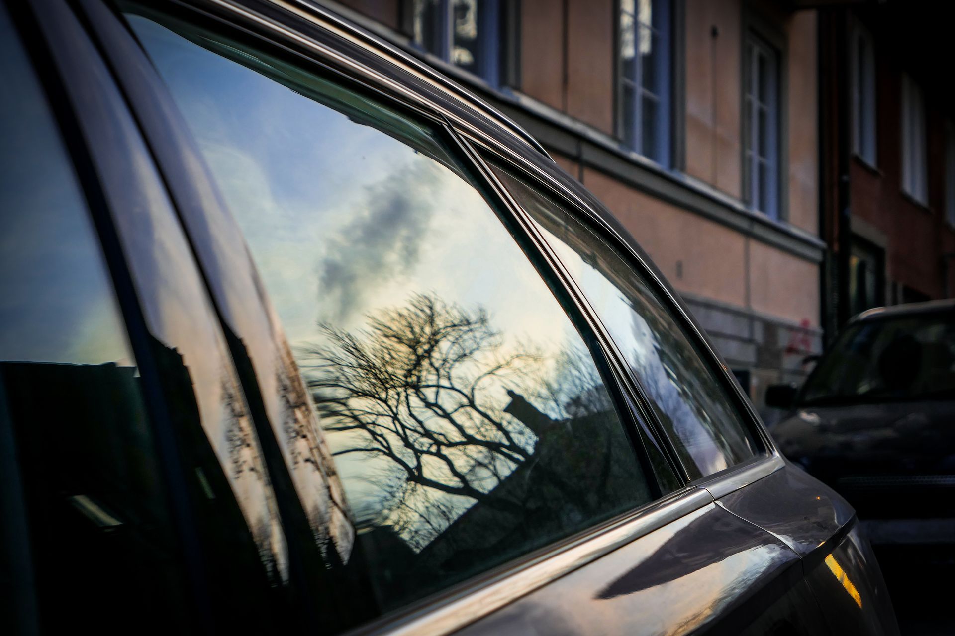 Reflections of a bare tree and cloudy sky in a car window beside a brick building