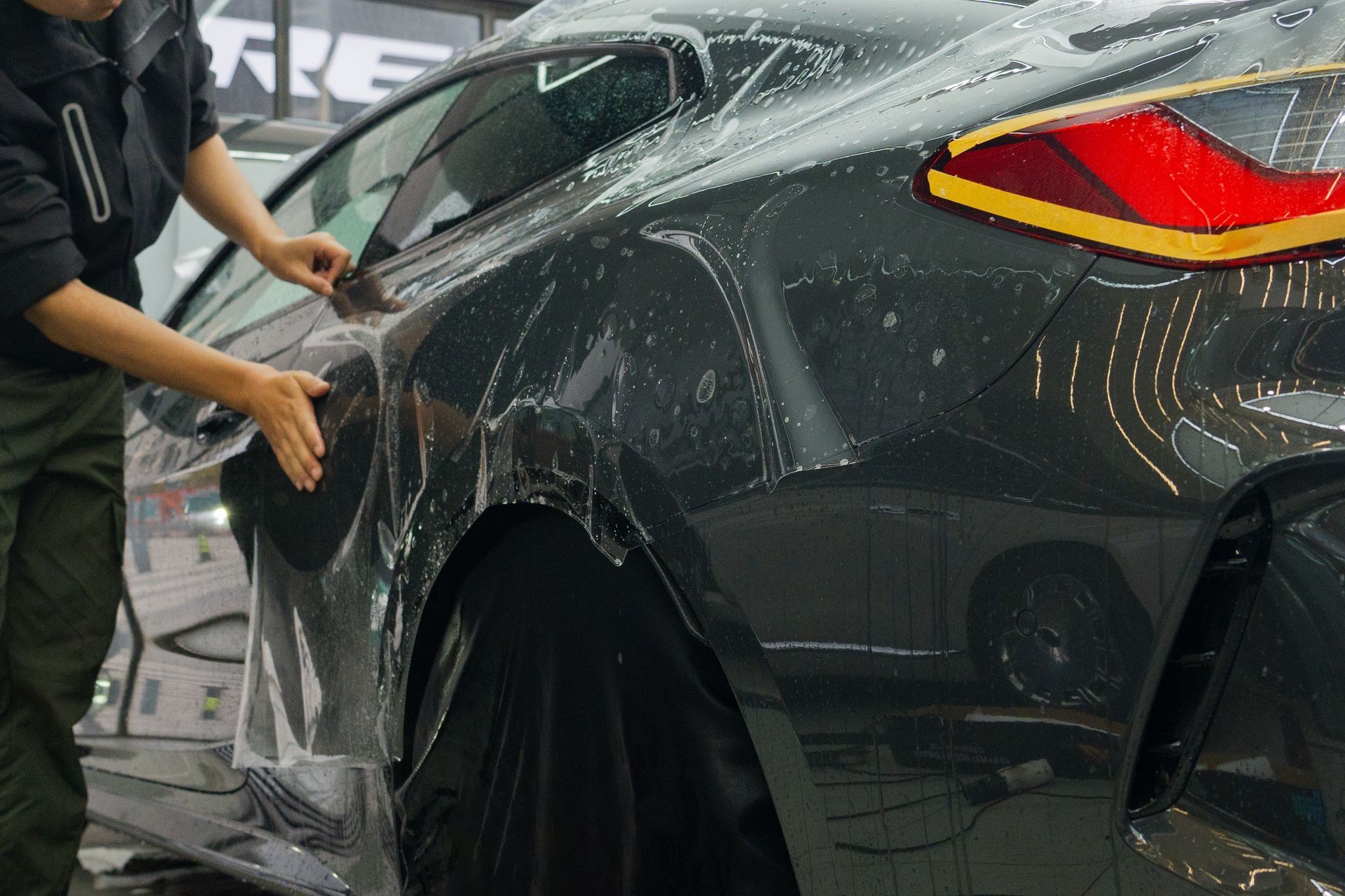 Person applying clear protective film to a black sports car in a workshop