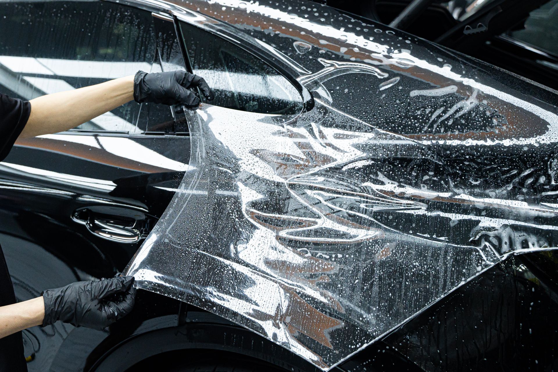 Gloved hands stretching clear protective film over a black car’s hood and fender