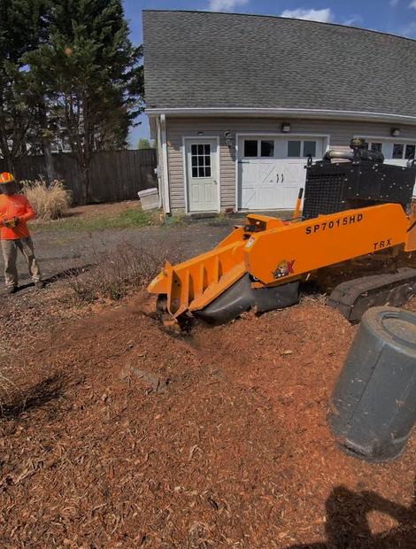 Orange construction machine on mulch-covered yard near a house, with a worker standing to the left.