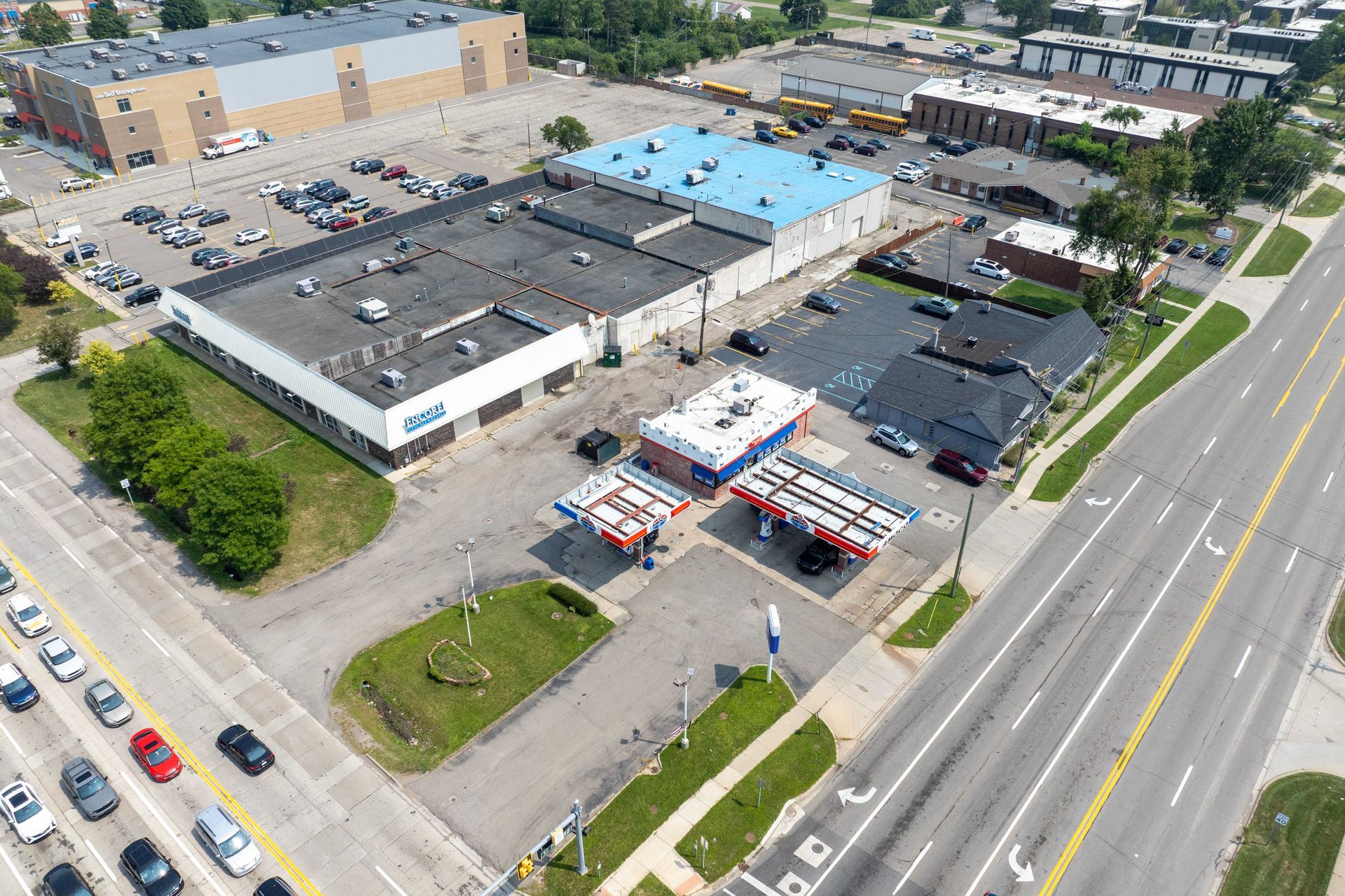 Aerial view: commercial buildings, gas station, busy road, parking lots, blue sky, cars.