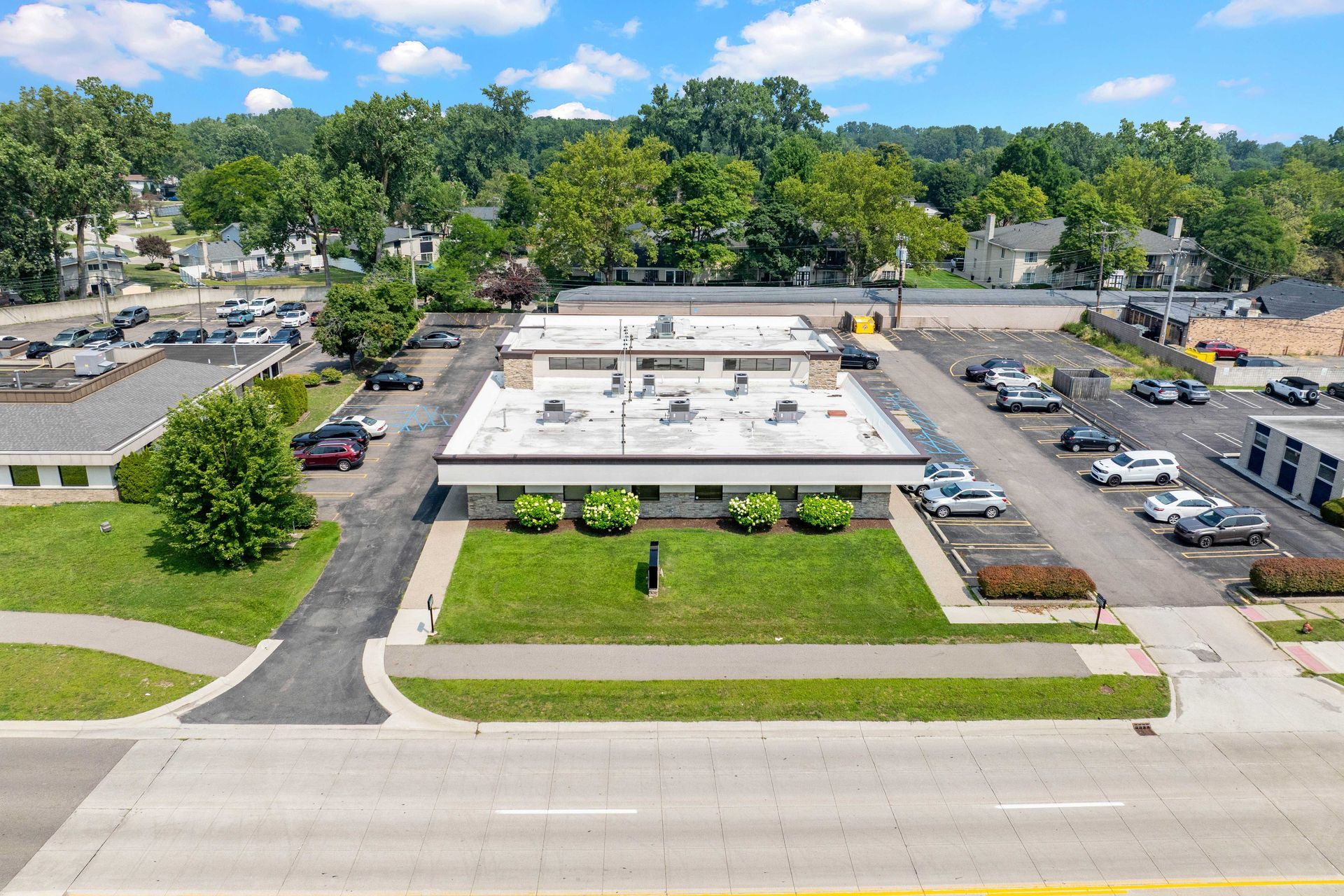 Aerial view of a commercial building with a flat roof, green lawn, and parked cars, surrounded by trees.