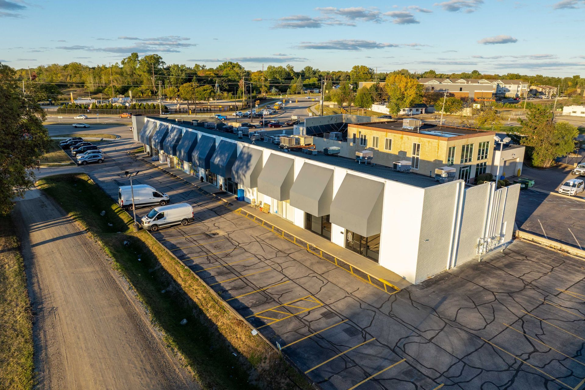 Aerial view of a long, gray commercial building with awnings; parking lot and road in front.