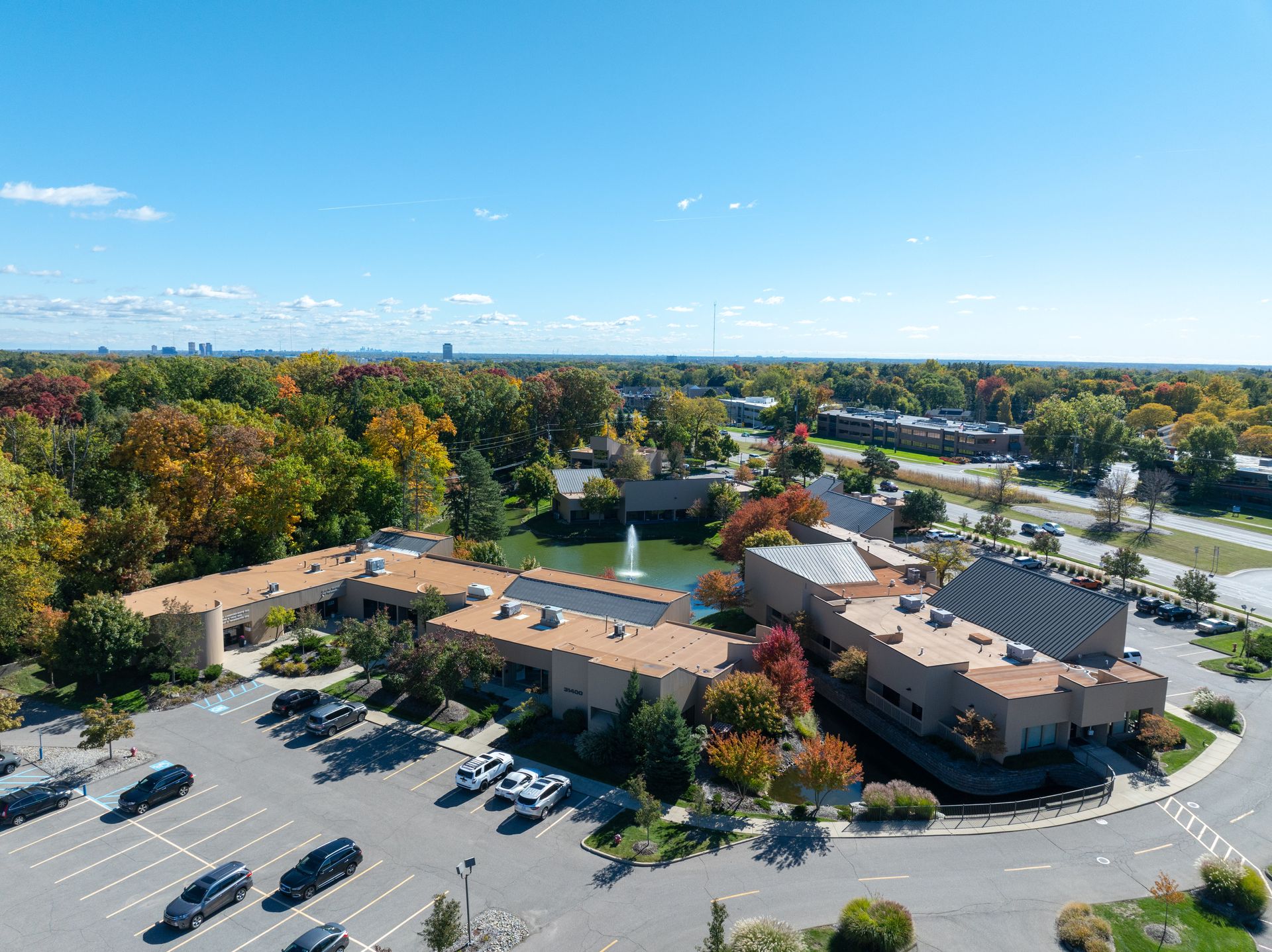 Aerial view of a light-colored building with a pond, surrounded by trees and a parking lot under a blue sky.