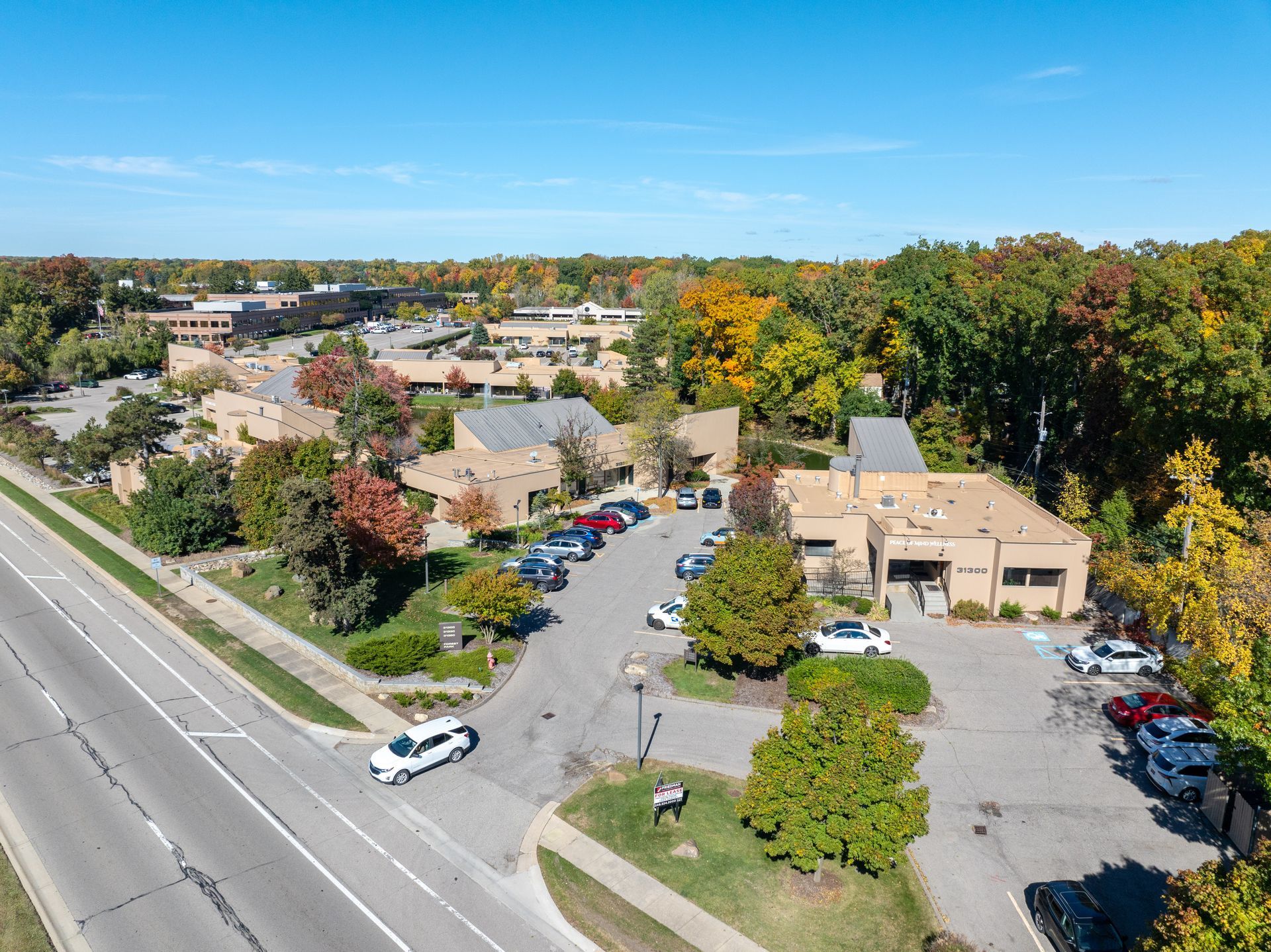 Aerial view of buildings, a road, and parked cars, with colorful autumn trees under a blue sky.