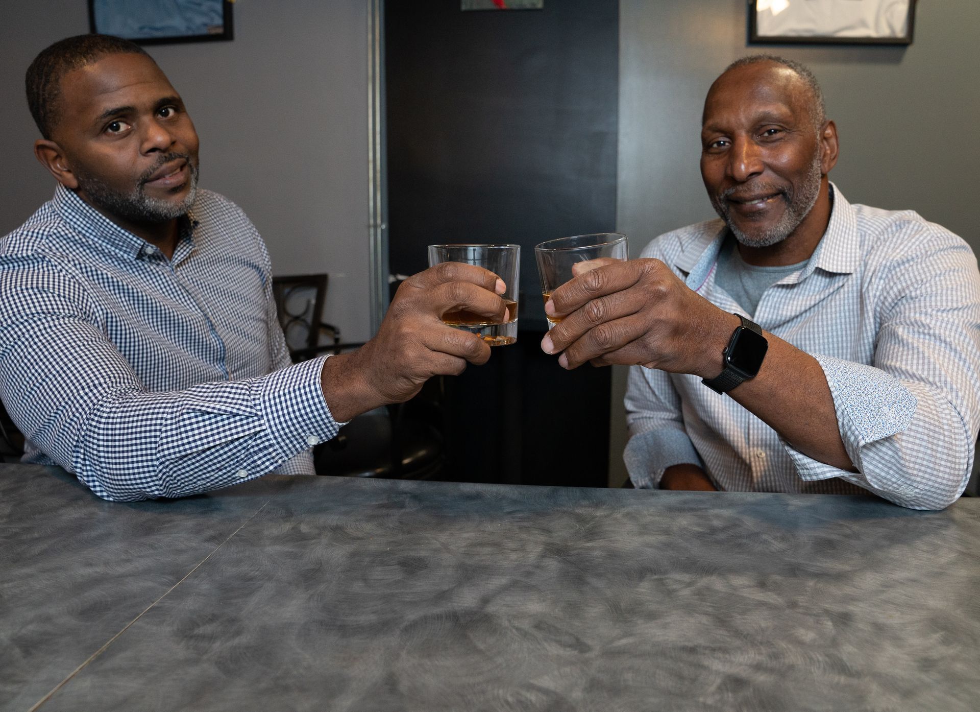 Two men are sitting at a table toasting with glasses of whiskey.