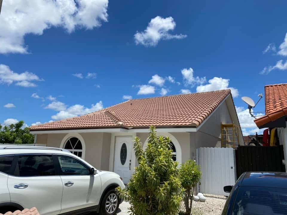 Single-story house with terracotta roof under a bright blue sky, with parked white and dark cars in front