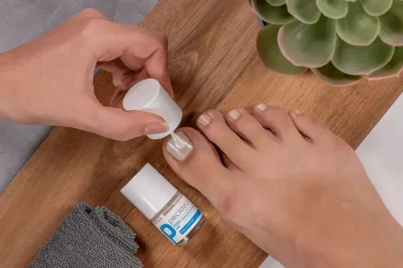 Person applying clear nail polish to toes on a wooden surface, with a bottle of polish and a small towel visible.