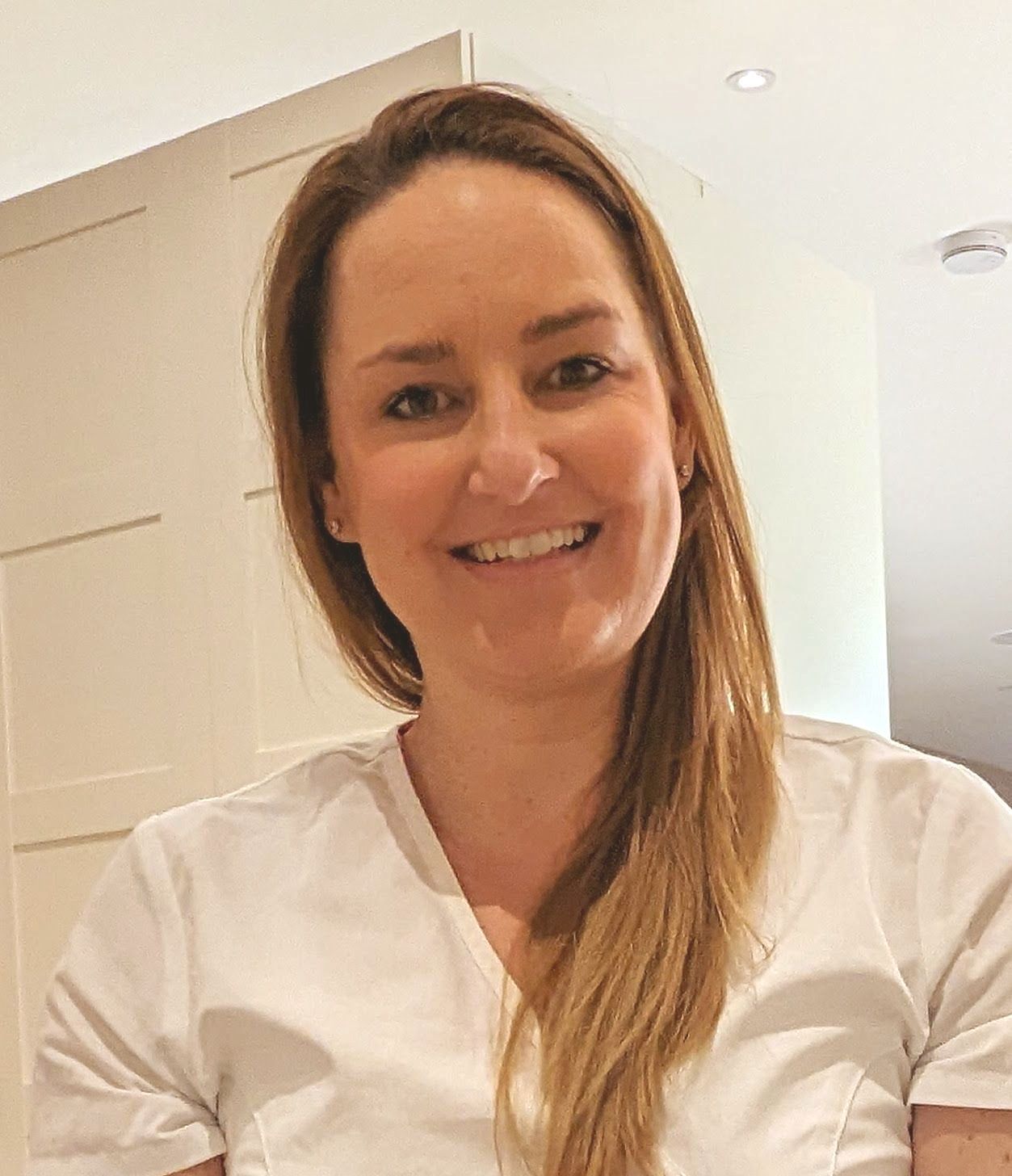 Woman with long, light brown hair smiles, wearing a white shirt; indoor setting.