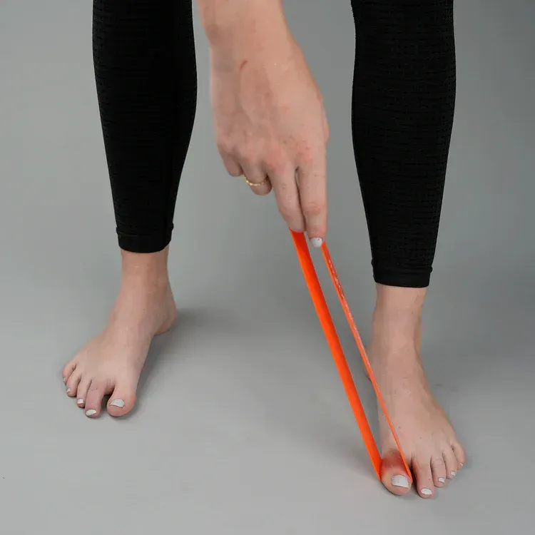 Person holding an orange resistance band, stretching foot and toes while standing on a gray surface.