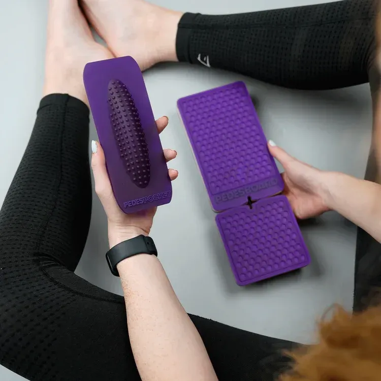 Person holding two purple foot massagers, with feet in the background.