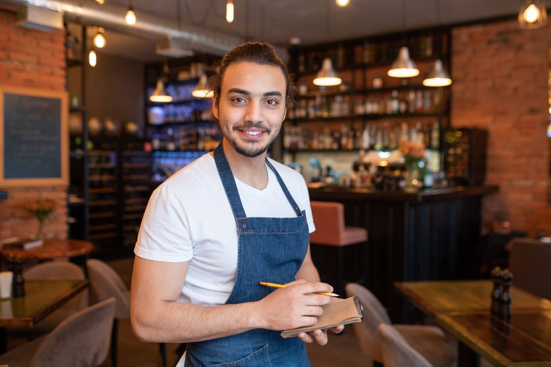 Barista sonriente con delantal vaquero de pie en el interior de una cafetería cálidamente iluminada.