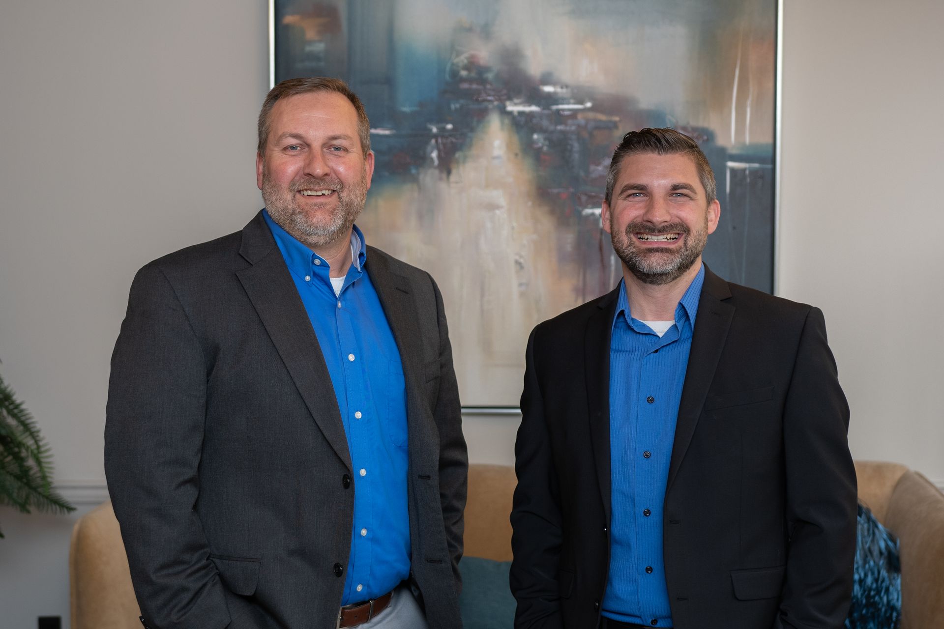 Two men in suits and blue shirts are standing next to each other in front of a painting.