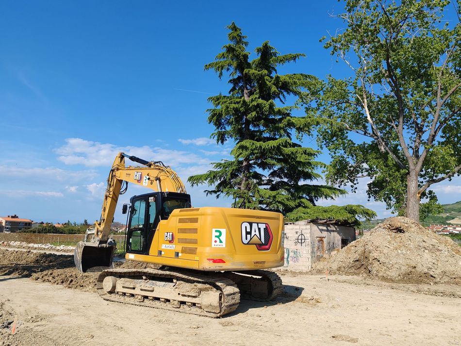 Yellow CAT excavator on a construction site beside dirt piles and trees under a blue sky
