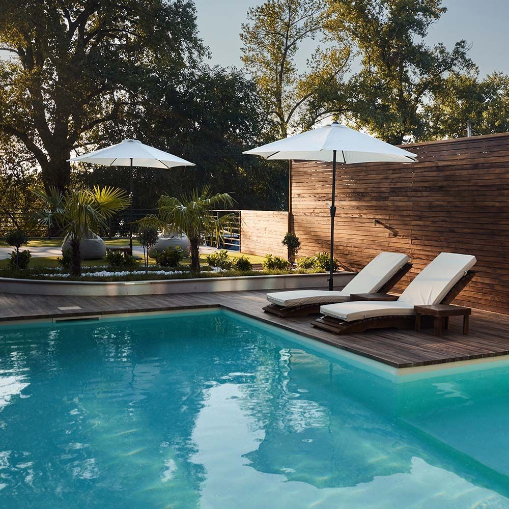 Backyard pool with two lounge chairs, white umbrellas, and trees beside a wooden fence